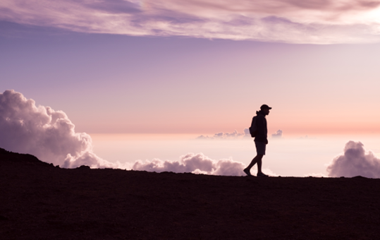 Silhouette of a person wearing a backpack walking against a pink and purple sunset sky.